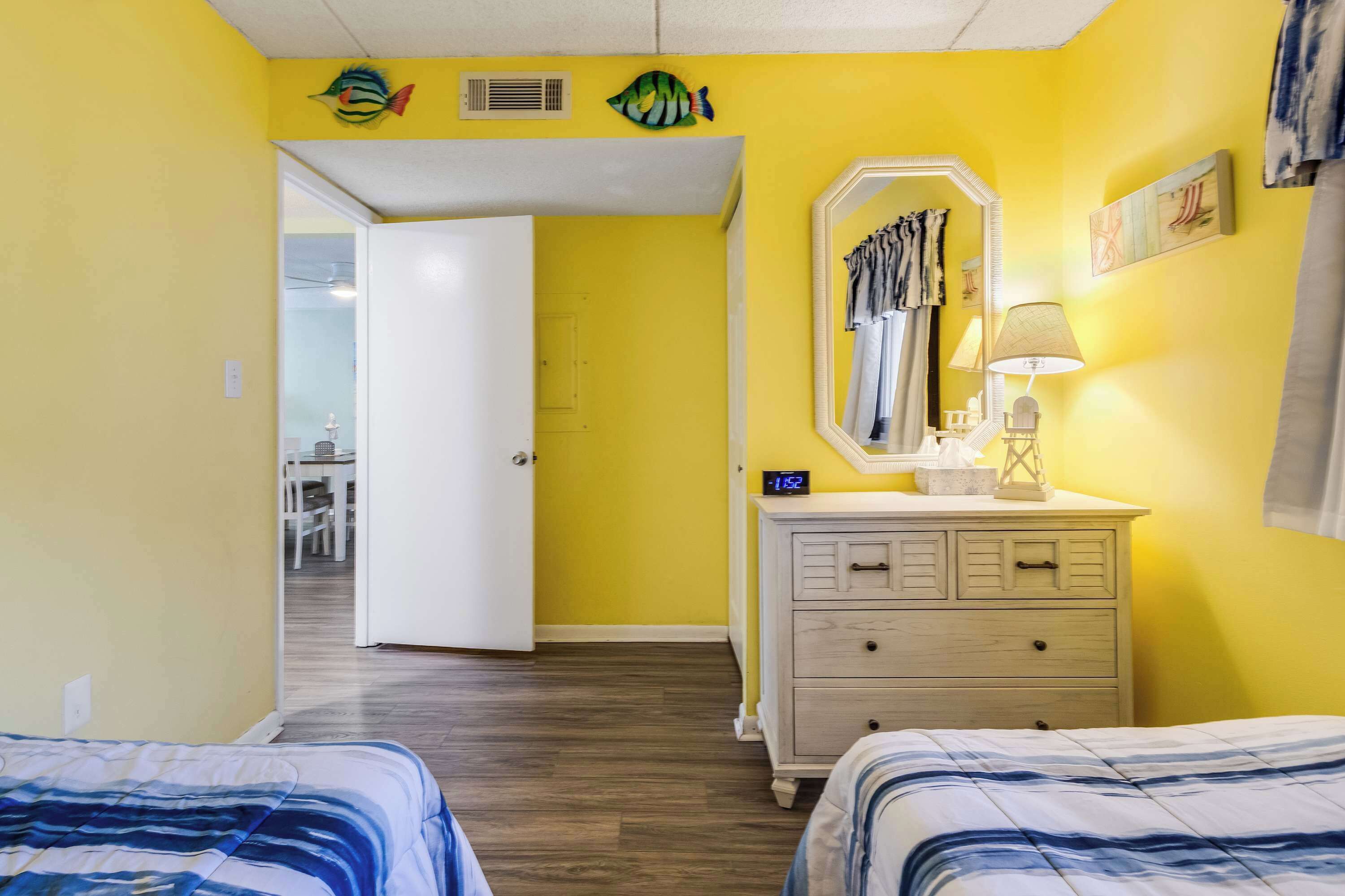 Una habitación con paredes amarillas, una puerta blanca, un aparador con espejo y dos camas con ropa de cama azul.