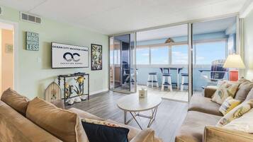 A spacious living room with a large sliding glass door leading to a balcony with a view of the ocean. There is a sofa, a coffee table, and a framed picture on the wall.