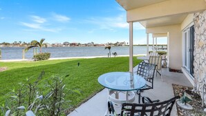 En overdækket terrasse med et glasbord og stole, med udsigt over vand og et boligområde i baggrunden.