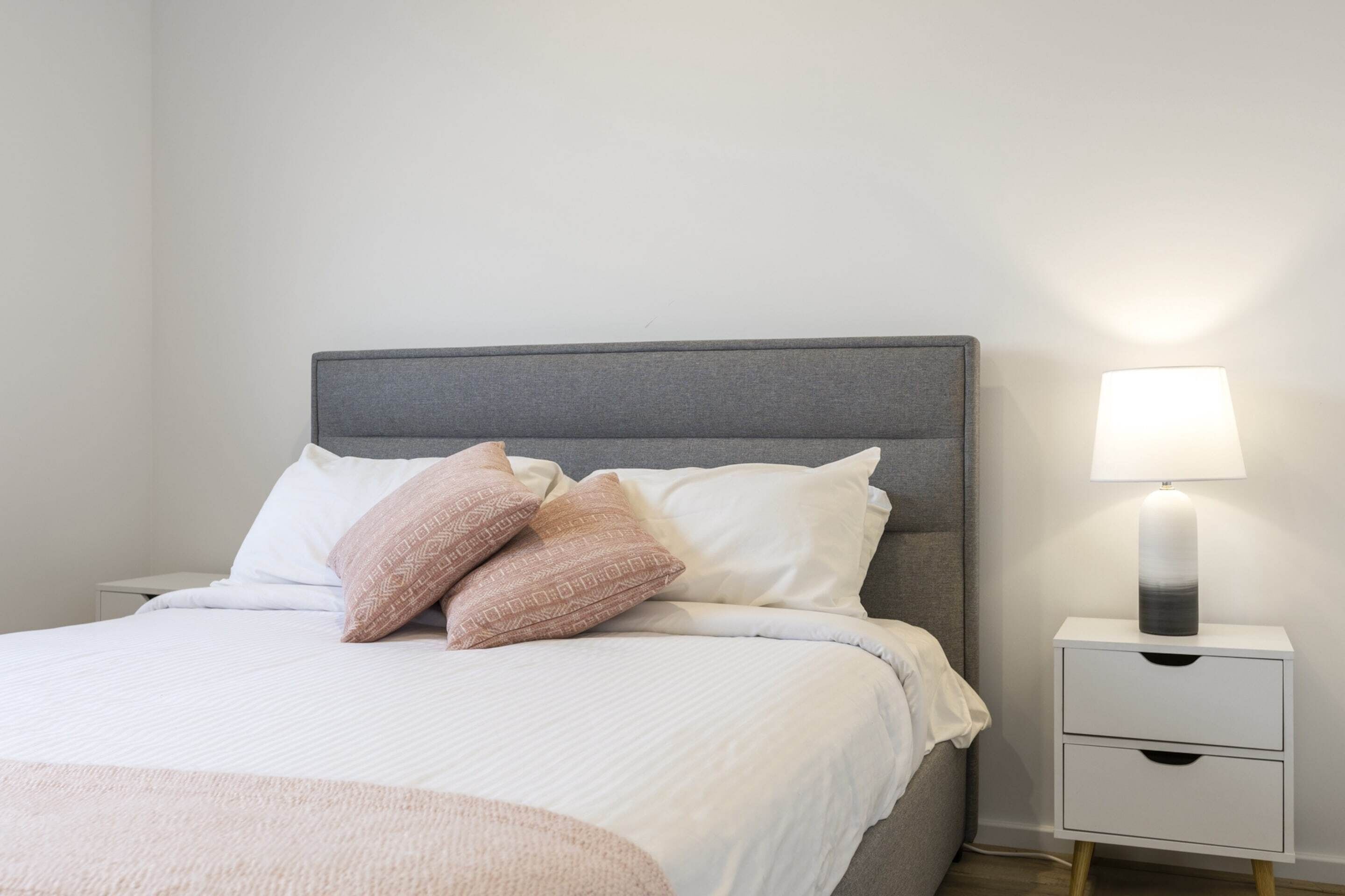 A neatly made bed with a grey headboard, white bedding, and two decorative pillows. There is a bedside table with a lamp and a drawer.
