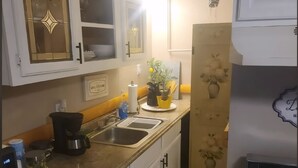 Keittiössä on valkoiset kaapit, liesi, pesuallas ja työtaso, jonka päällä on erilaisia esineitä.
