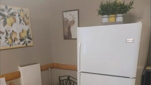 Kompakti keittiö, jossa on valkoinen jääkaappi, puinen ruokapöytä, jonka päällä on keltainen kulho, sekä kuviollinen koristetyynyillä varustettu tuoli.