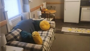 Kompakti olohuone, jossa on ruutukuviollinen sohva, pieni ruokapöytä, jääkaappi ja ikkuna, jossa on sälekaihtimet.