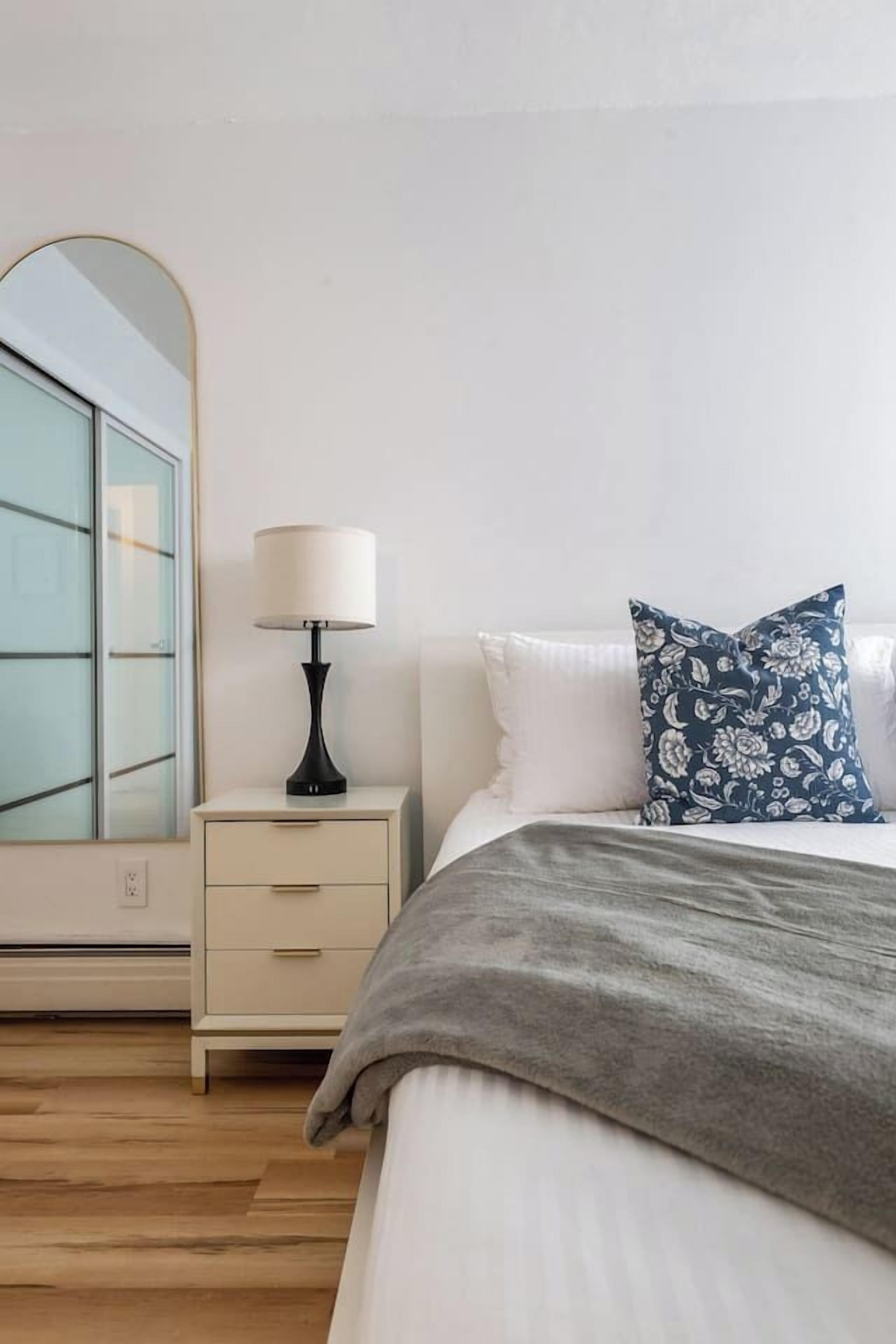 A bedroom with a bed, a nightstand with a lamp, a dresser, and a large mirror.