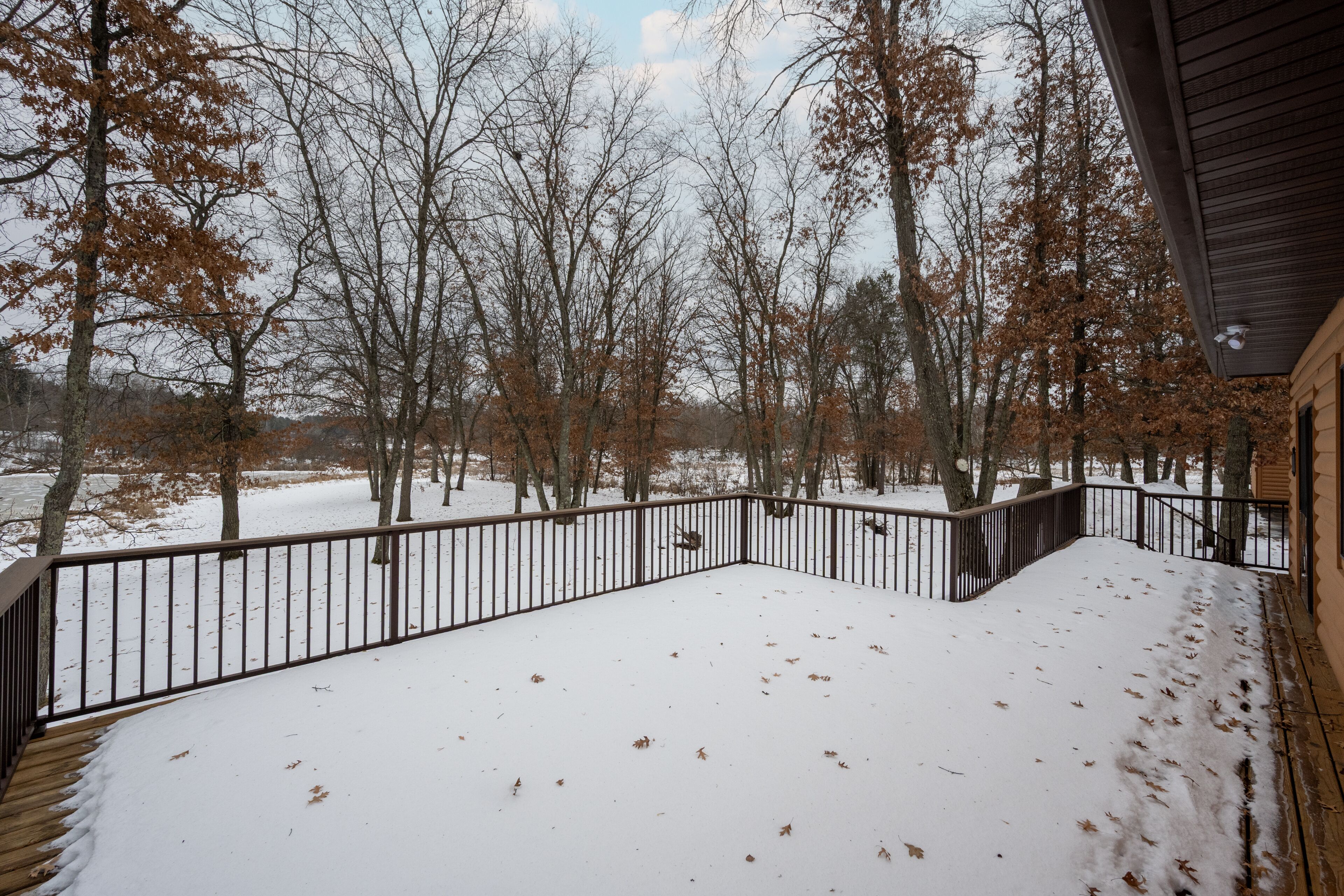 A snow-covered deck with a metal railing, surrounded by leafless trees and a wooden building.