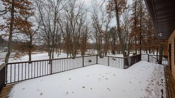 A snow-covered deck with a metal railing, surrounded by leafless trees and a wooden building.
