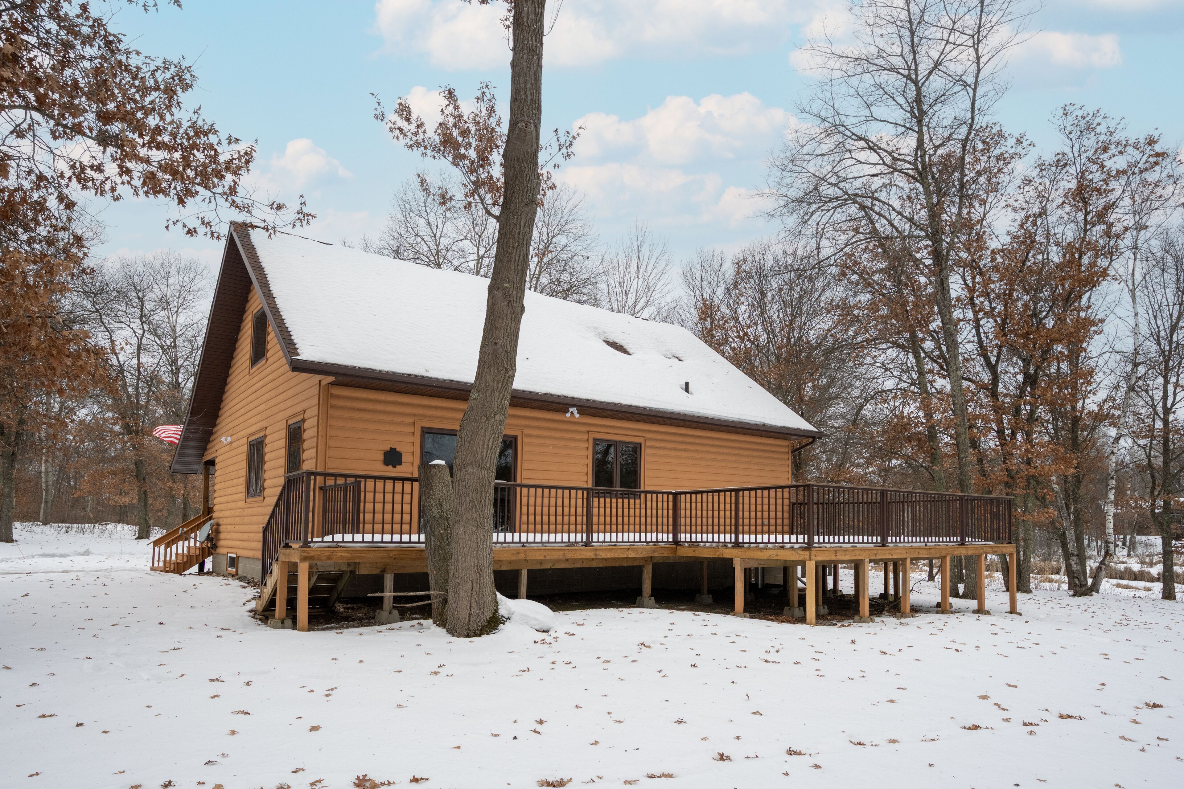 A wooden cabin with a deck and a snow-covered roof, surrounded by trees.