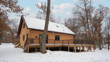 A wooden cabin with a deck and a snow-covered roof, surrounded by trees.