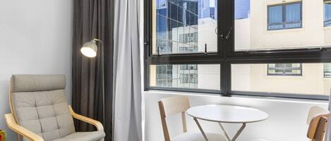 A room with a grey upholstered chair, a small round table, and a floor lamp. A window offers a view of buildings outside.
