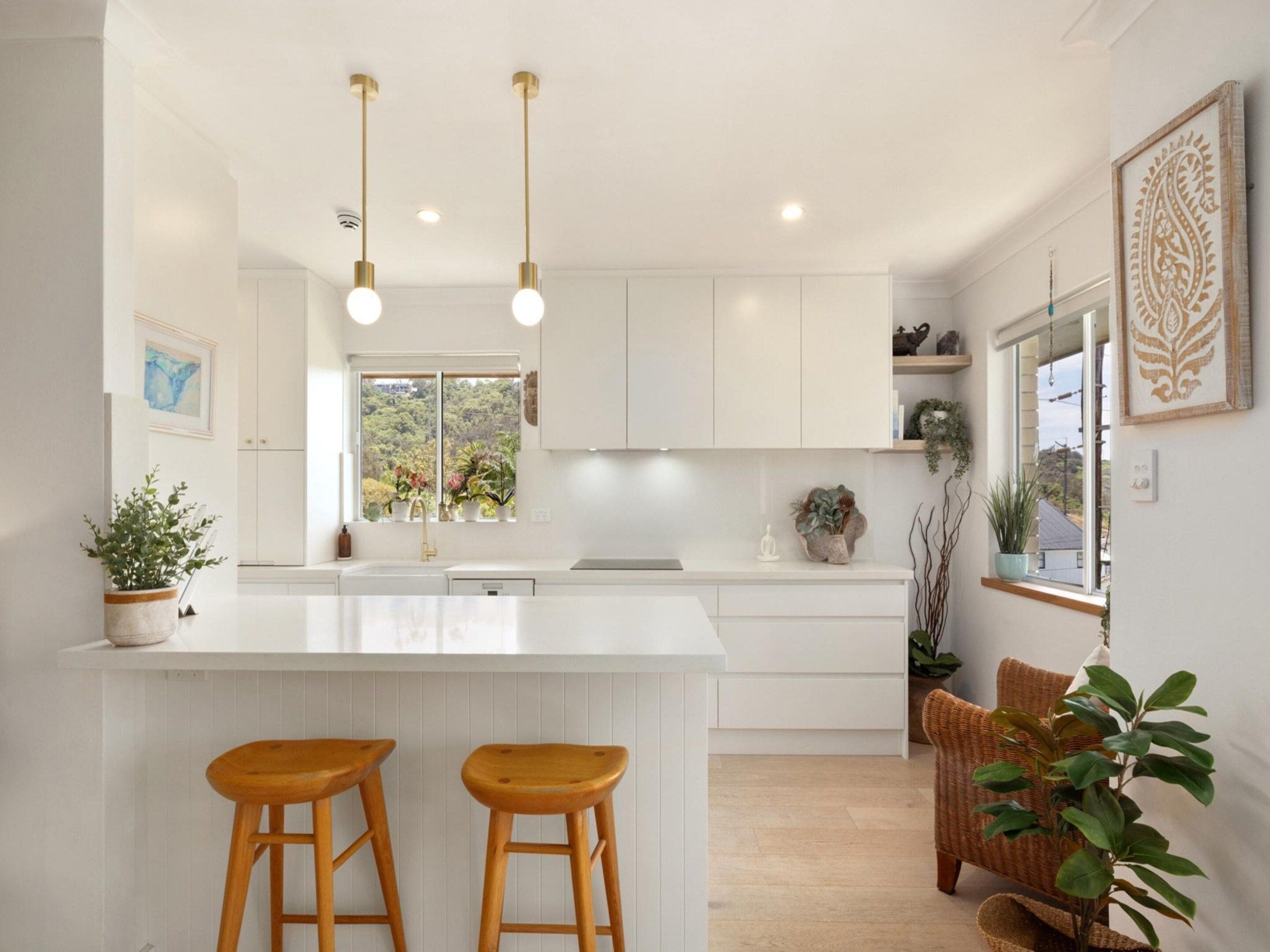 A modern kitchen with white cabinetry, a central island with stools, pendant lighting, and a view of greenery outside.