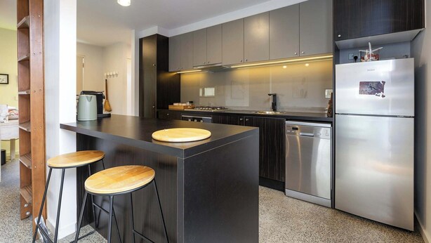 A modern kitchen with dark cabinetry, a stainless steel refrigerator, a dishwasher, and a central island with stools. - Spacious 1-bed Townhouse in Norlane (Geelong)