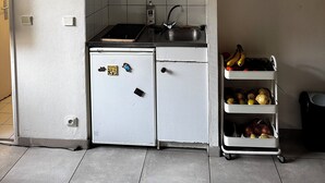 Eine kleine Küche mit weißen Schränken, einem schwarzen Herd und einer Spüle. Auf der Arbeitsfläche steht ein kleiner Kühlschrank und ein Obstkorb.