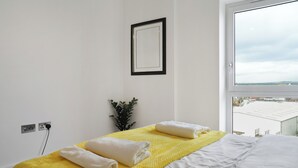Cama bem arrumada com lençóis brancos e manta amarela, uma pequena planta em vaso e uma janela com vista para edifícios.