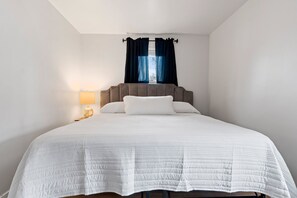 Un lit bien fait, recouvert d’une couette blanche, une table de chevet en bois avec une lampe, et une fenêtre avec des rideaux foncés.