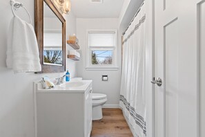 Une salle de bain avec un lavabo blanc, des toilettes et un rideau de douche. Il y a une fenêtre au-dessus des toilettes et une serviette accrochée au mur.