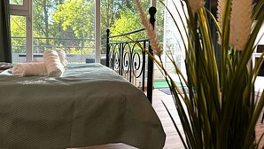 A bedroom with a bed, a view of trees through large windows, and a railing outside.