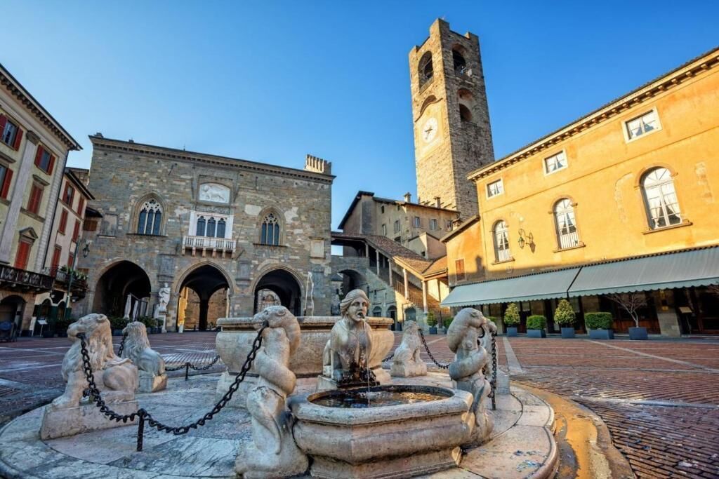 A historic square with a fountain, stone buildings, and a tall tower.