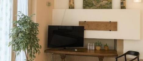 A living room with a flat-screen TV on a wooden stand, a potted plant, a dining table with chairs, and a shelf with decorative items.