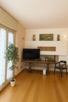 Un soggiorno con una televisione a schermo piatto su un mobile in legno, una pianta in vaso, un tavolo da pranzo con sedie e una mensola con oggetti decorativi.