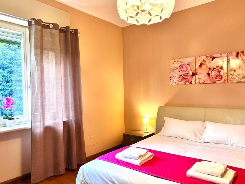 Una camera d'albergo con un letto, un comodino, una lampada, una finestra con tende e un quadro floreale appeso al muro.