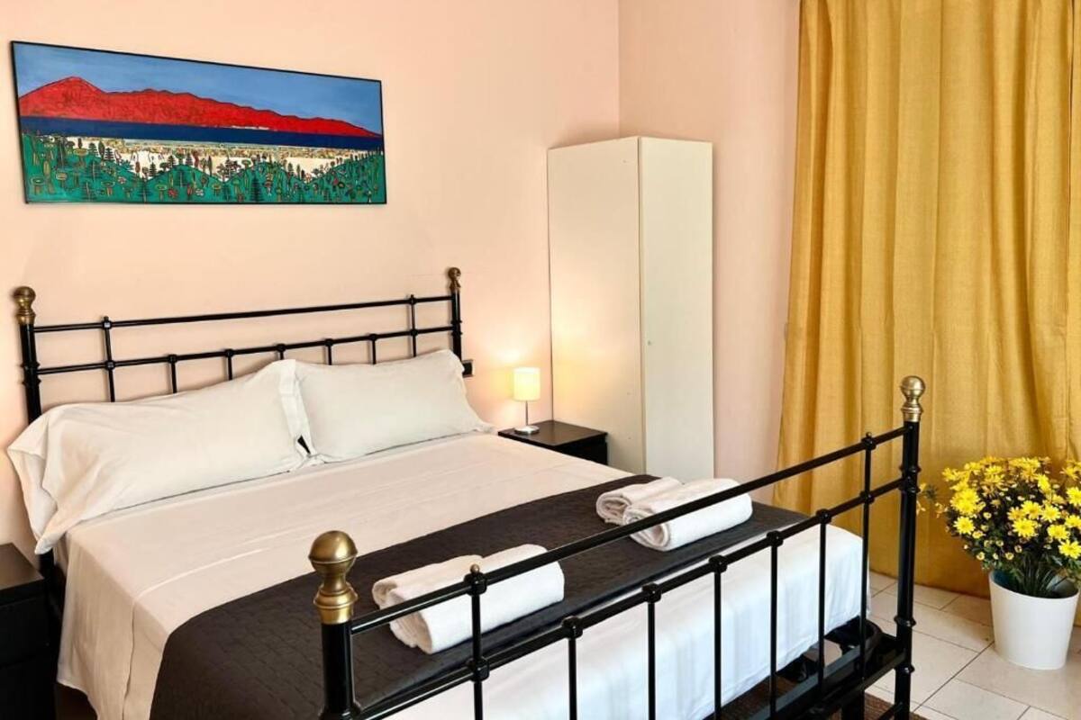 A neatly made bed with a headboard, a bedside table with a lamp, a wardrobe, and a painting of a landscape on the wall.