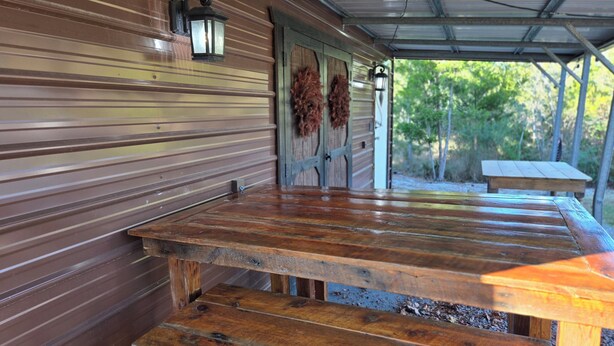 Un espacio de estar al aire libre con techo, bancos de madera y una mesa, una puerta metálica decorada con coronas y un farol en la pared.