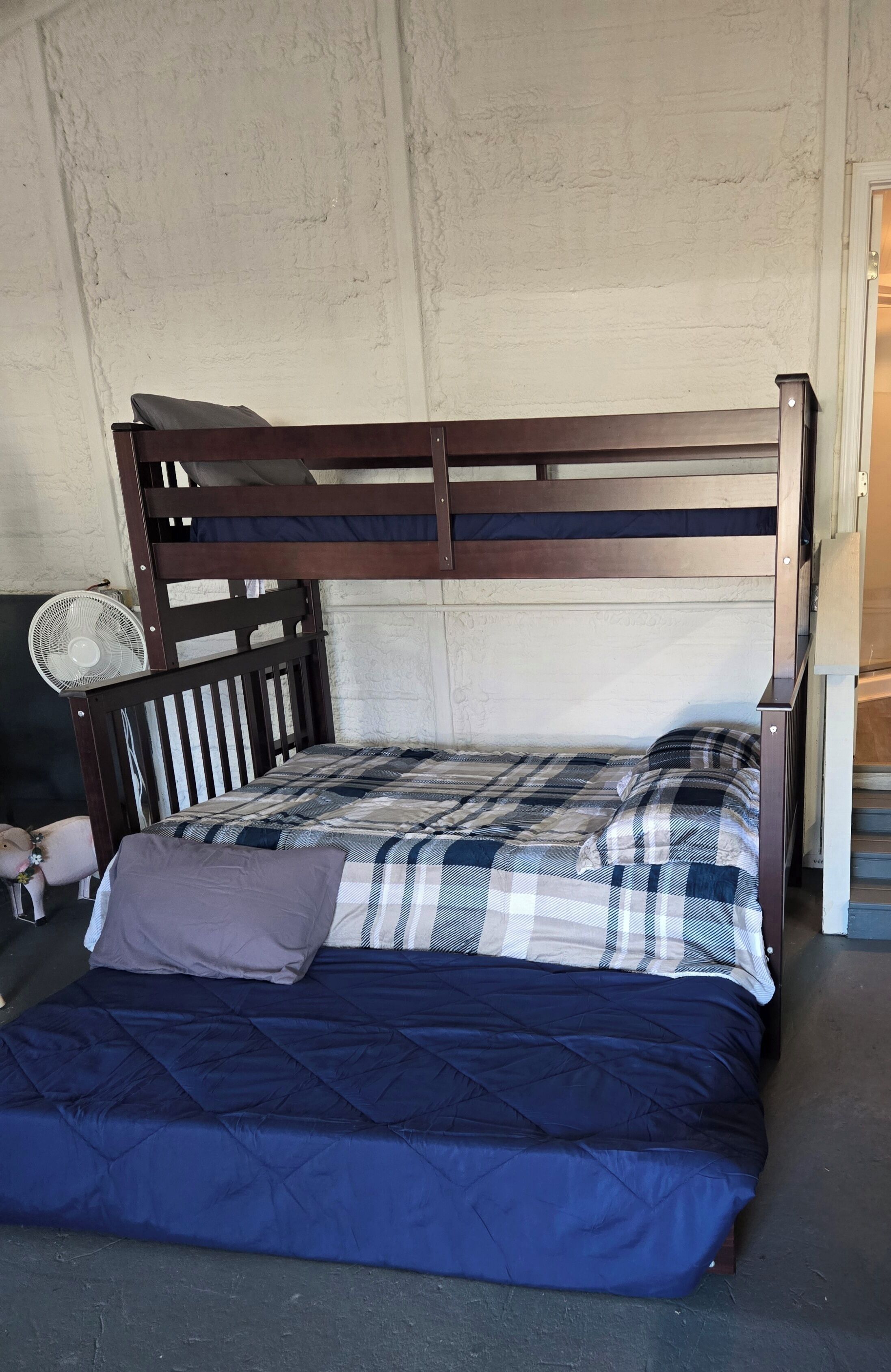 Un lit superposé avec une couverture à carreaux et des oreillers, un ventilateur sur pied et un petit objet décoratif posé sur le sol.