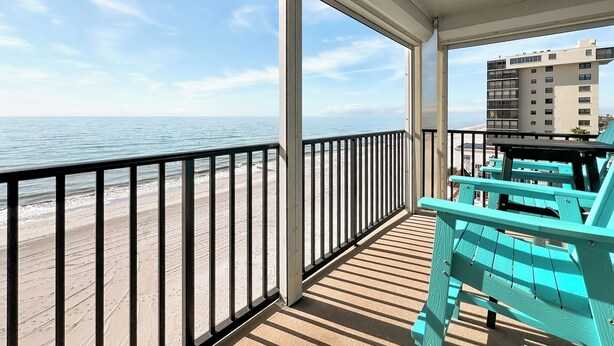A balcony with a view of the beach and ocean, wooden deck, turquoise chairs, and a building in the background. - “Sun, Sand & Spectacular Views Family-Friendly Madeira Beach Condo-Corner Unit (Madeira Beach)