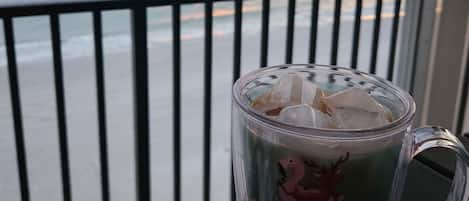 A glass mug with ice cubes on a balcony overlooking the ocean at sunset.