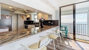 A modern kitchen with a marble countertop, white bar stools, and a view of the ocean through a sliding glass door. - “Sun, Sand & Spectacular Views Family-Friendly Madeira Beach Condo-Corner Unit (Madeira Beach)