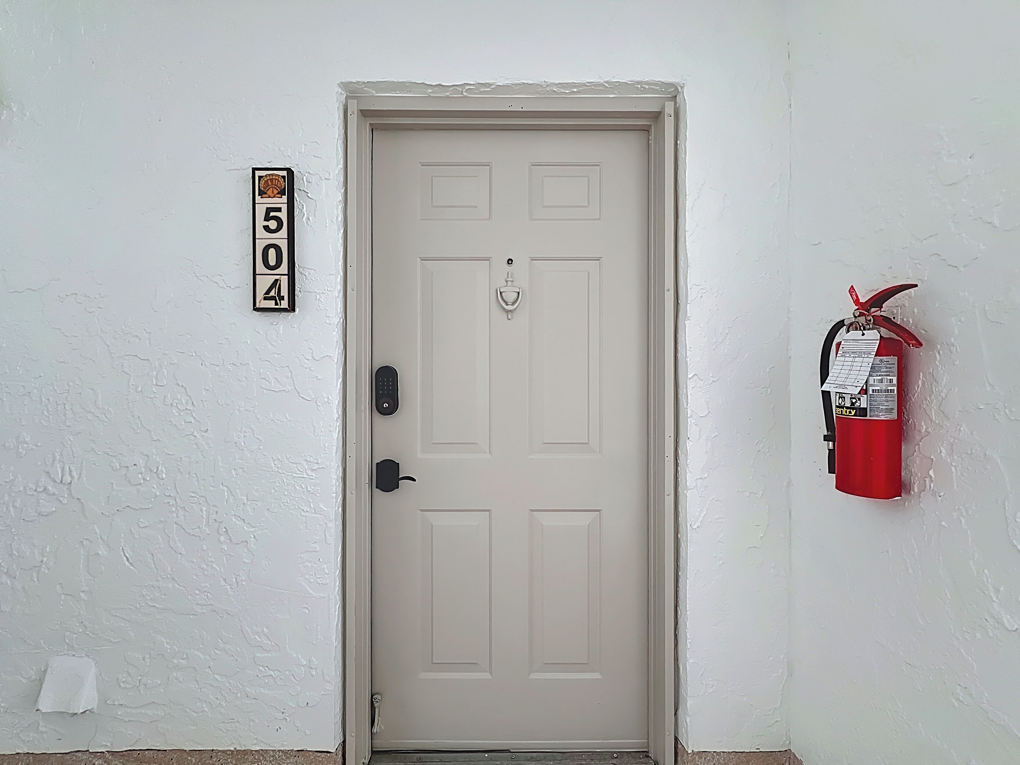 Una porta bianca con serratura a combinazione, un estintore fissato al muro sulla destra e un numero civico sopra la porta.