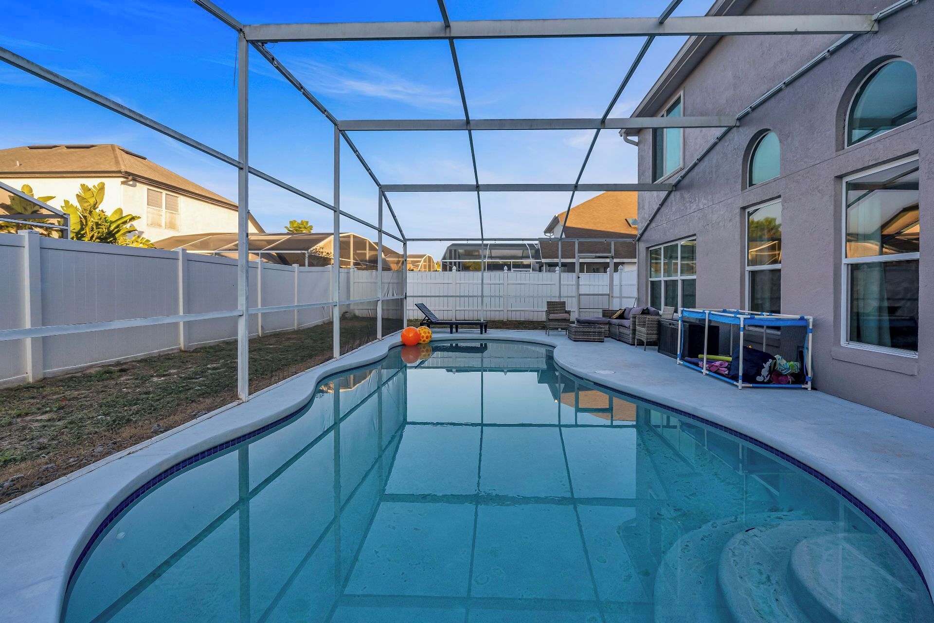 A swimming pool with a covered area, surrounded by a white fence and a house with large windows.