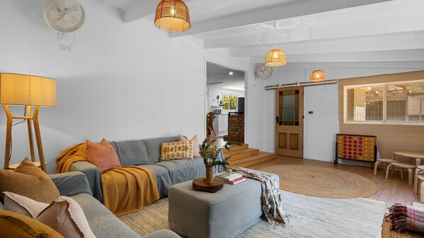 A modern living room with a sofa, ottoman, and a dining area with a table and chairs. There is a staircase leading to an upper level.