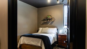 Een slaapkamer met een bed, nachtkastje, dressoir, een geometrisch wandplankje met planten, en een raam.