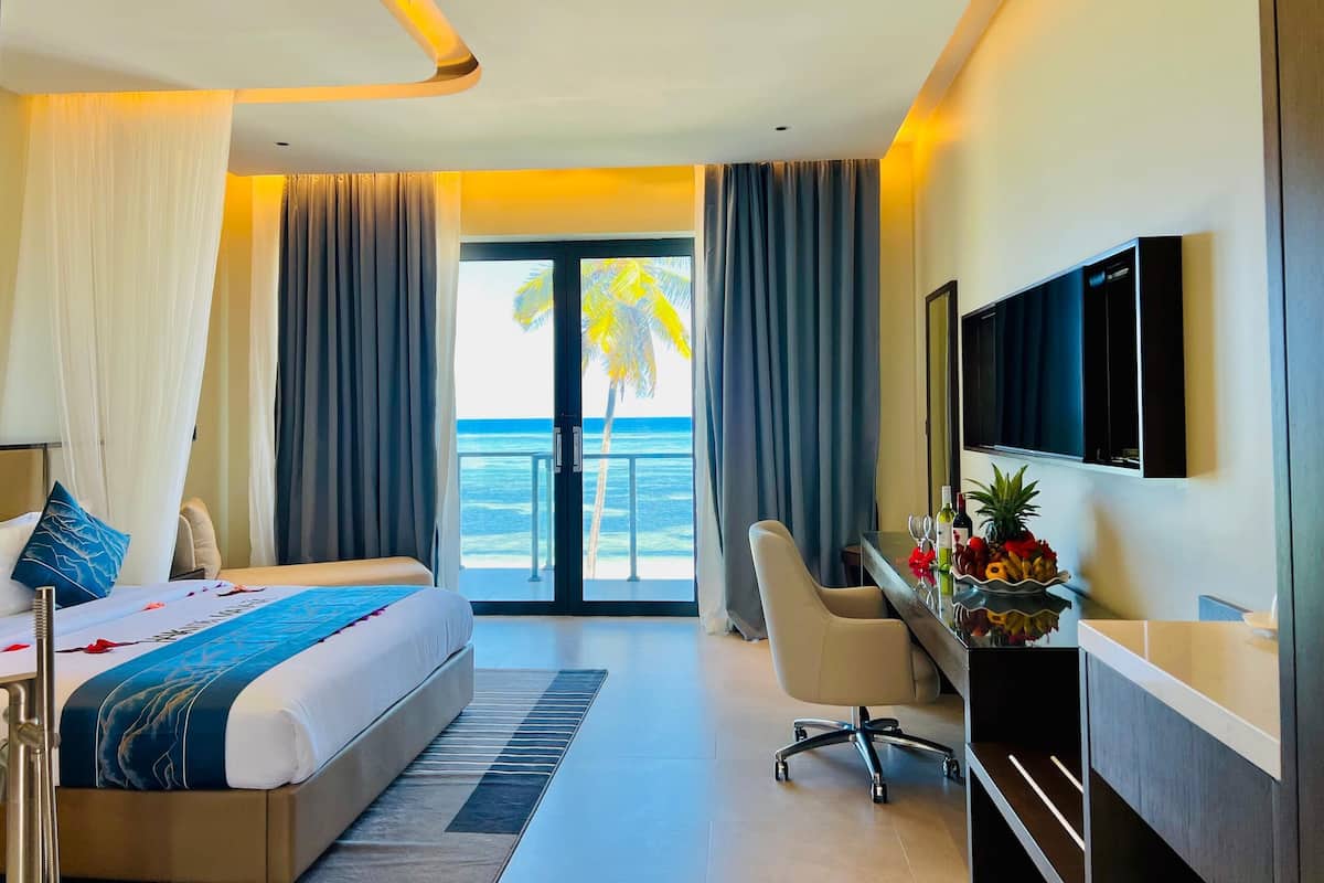 A hotel room with a bed, desk, and TV. The room has a view of the ocean and palm trees.