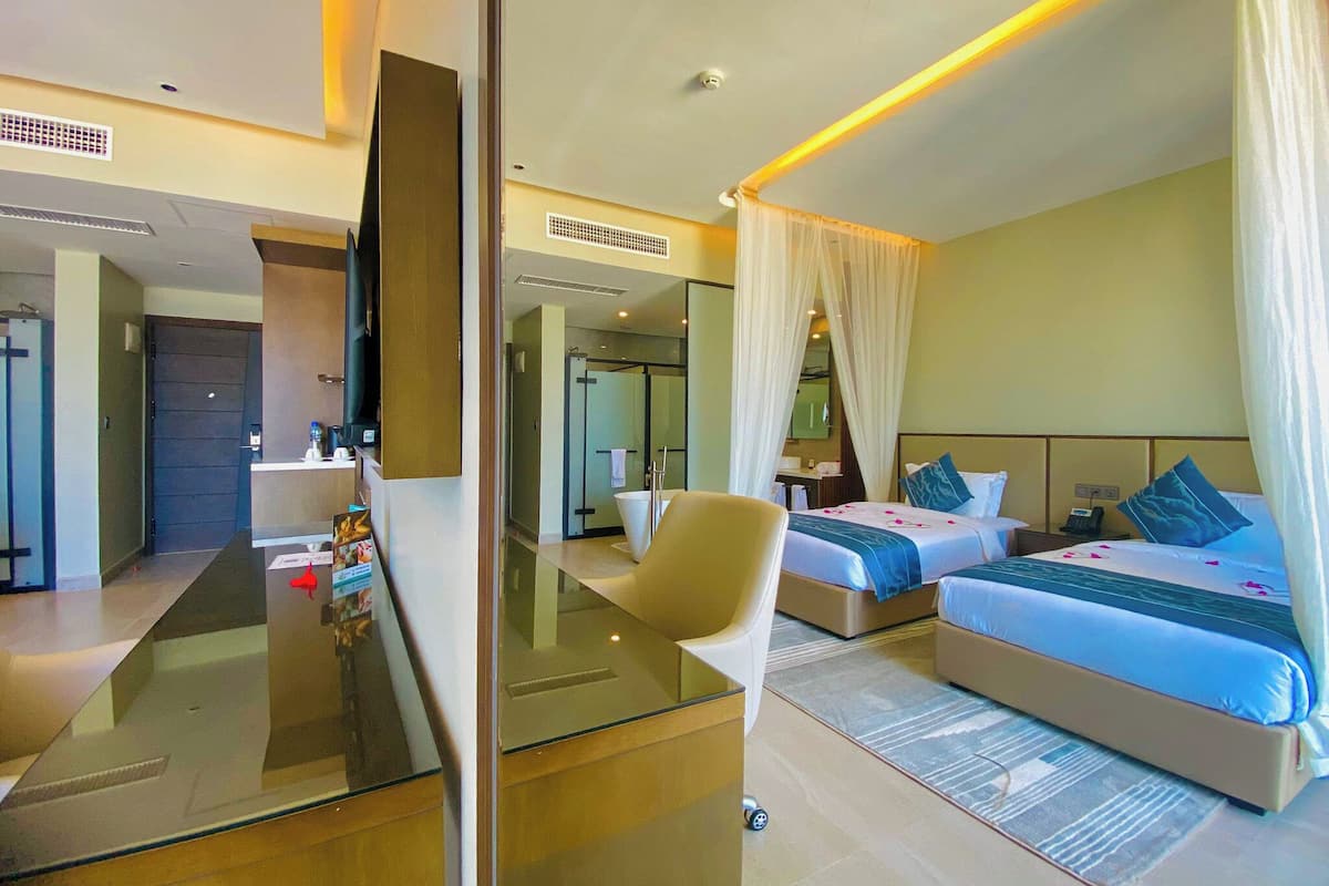 A modern hotel room with a glass partition, two beds, a sitting area, and a bathroom visible through the glass.