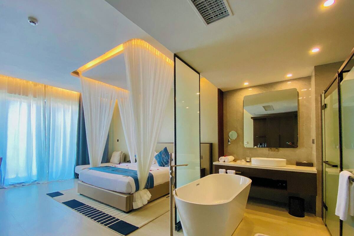 A modern hotel room with a large bed, a separate bathroom with a freestanding bathtub, and a large mirror above the sink.