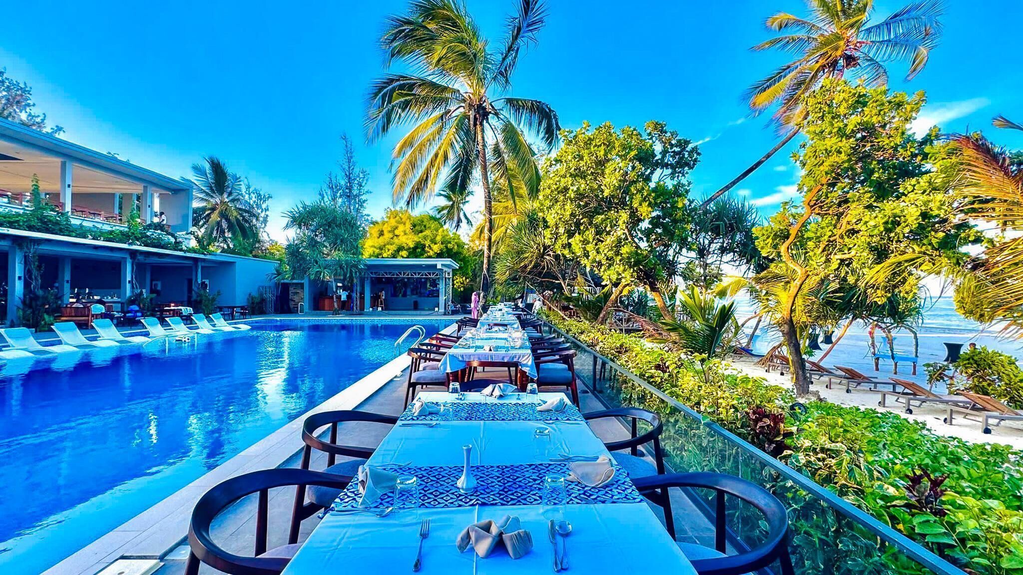 A poolside dining area with tables set for two, overlooking a swimming pool, surrounded by palm trees and lush greenery.