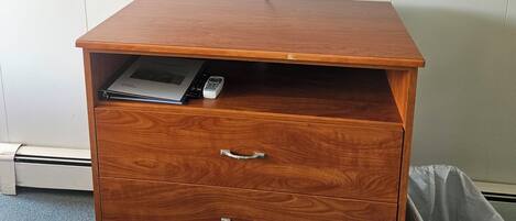 A room with a bed, a television mounted on the wall, a wooden cabinet with drawers, and a trash bin.