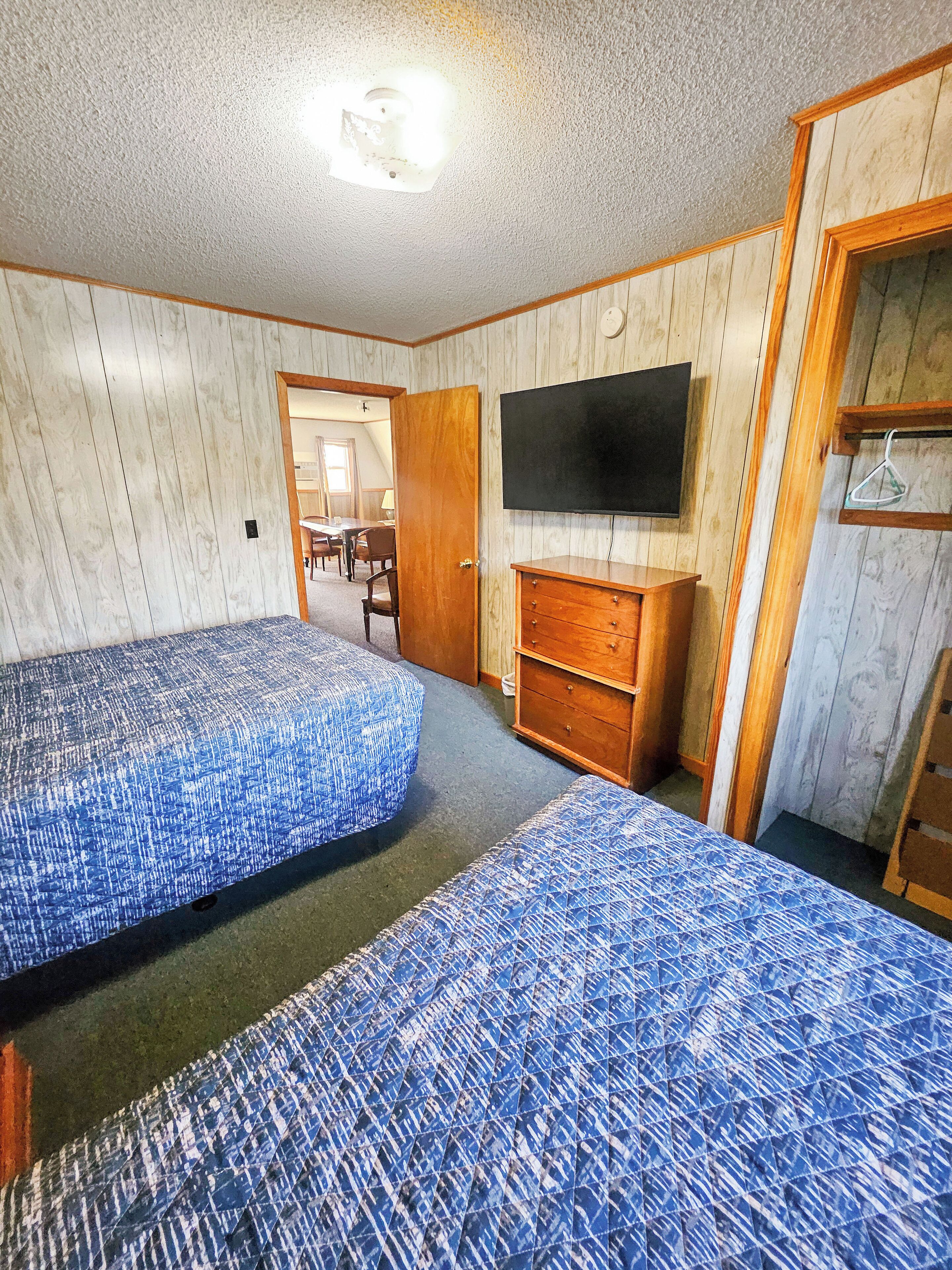 A room with two beds, a TV, a wooden dresser, and a door leading to another room.