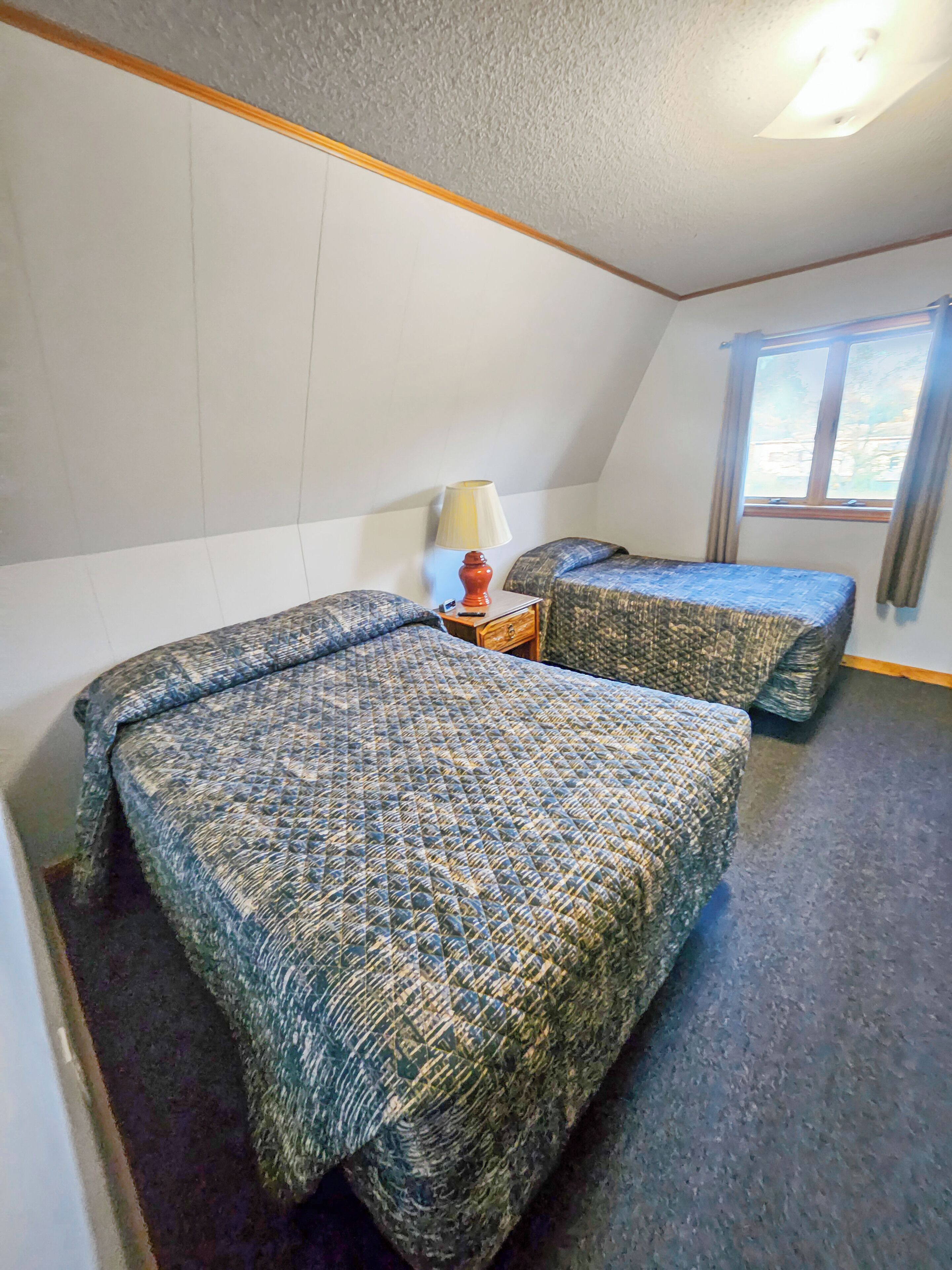 A room with two beds, a nightstand with a lamp, a window with curtains, and a ceiling light.