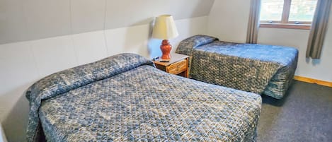 A room with two beds, a nightstand with a lamp, a window with curtains, and a ceiling light.
