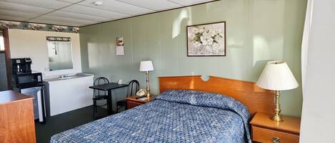 Hotelkamer met bed, nachtkastje, lamp, bureau, stoel, kitchenette en een schilderij aan de muur.