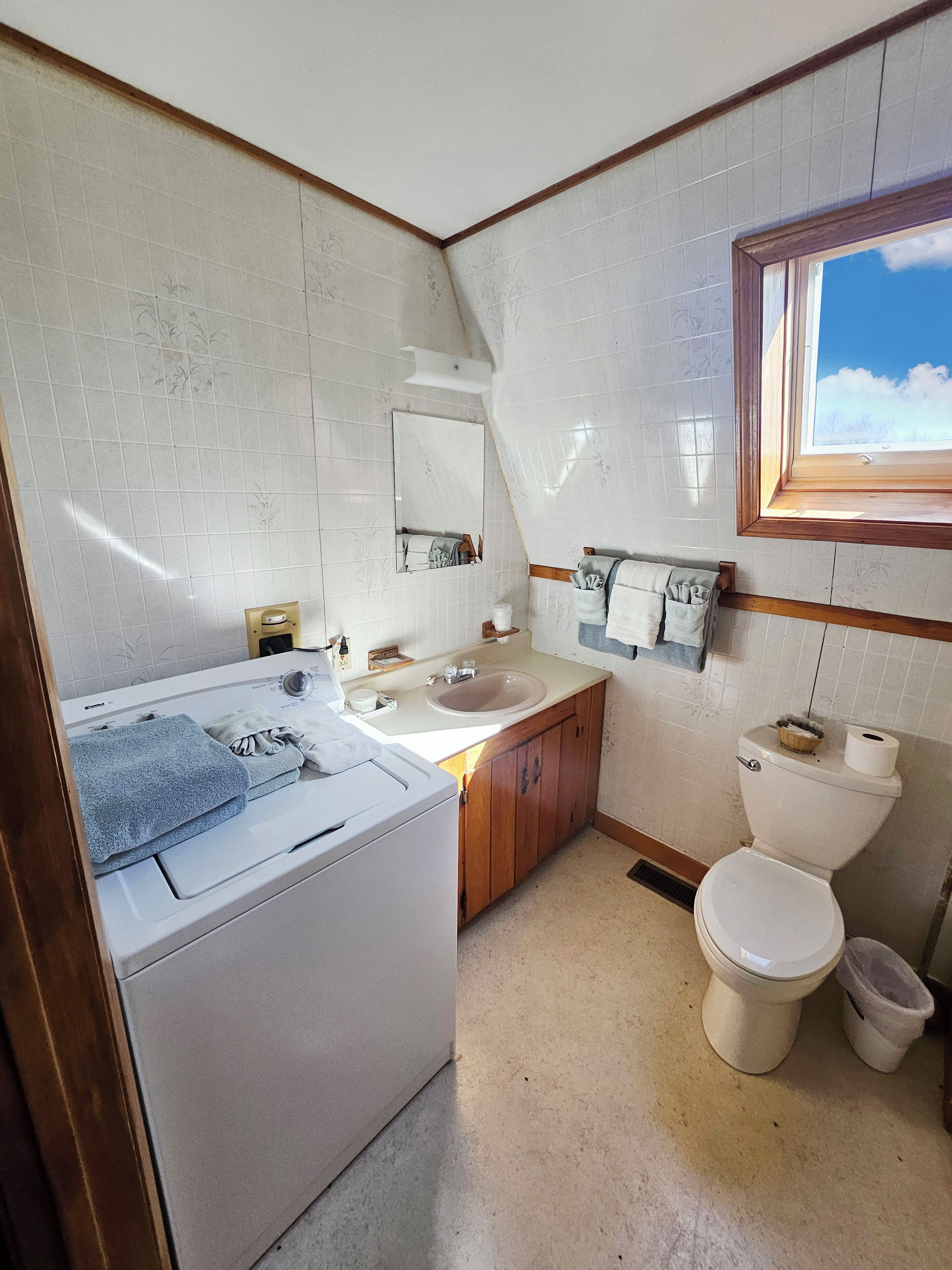 A compact bathroom with a washing machine, a sink, a toilet, and a window with a view of the sky.