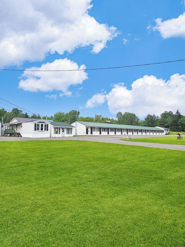A motel with a green roof and white exterior, located next to a grassy field under a clear blue sky. - Katahdin Valley Motel (Sherman Mills)