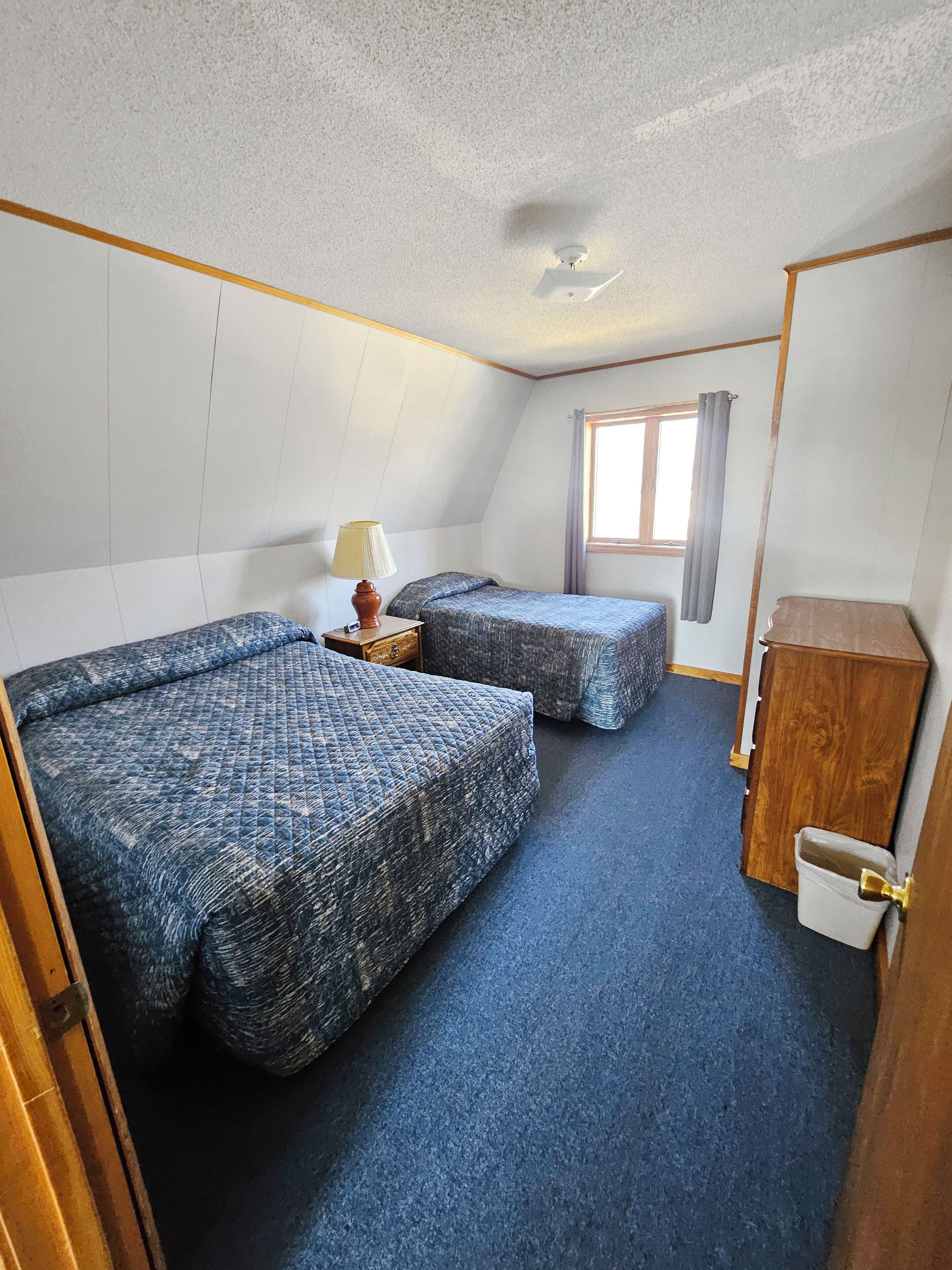 Une chambre avec deux lits, une petite table avec une lampe, une fenêtre avec des rideaux et un meuble en bois.