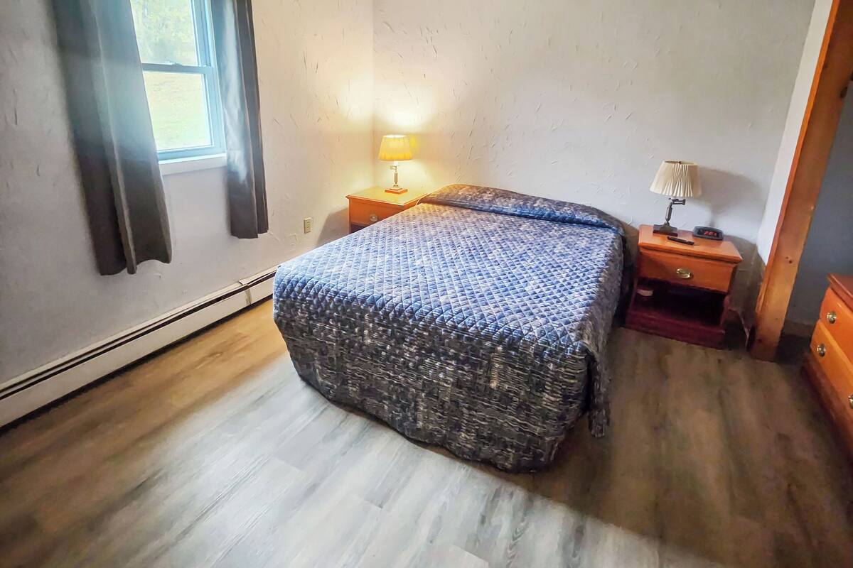 A bedroom with a bed, two bedside tables, a window with curtains, and a wooden floor.
