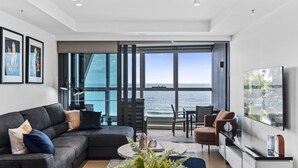 A modern living room with a large window overlooking the sea, a black sofa, a dining area, and a TV on the wall. - Luxury Beachfront 1-bed Apartment With Pool & Gym (Gold Coast)