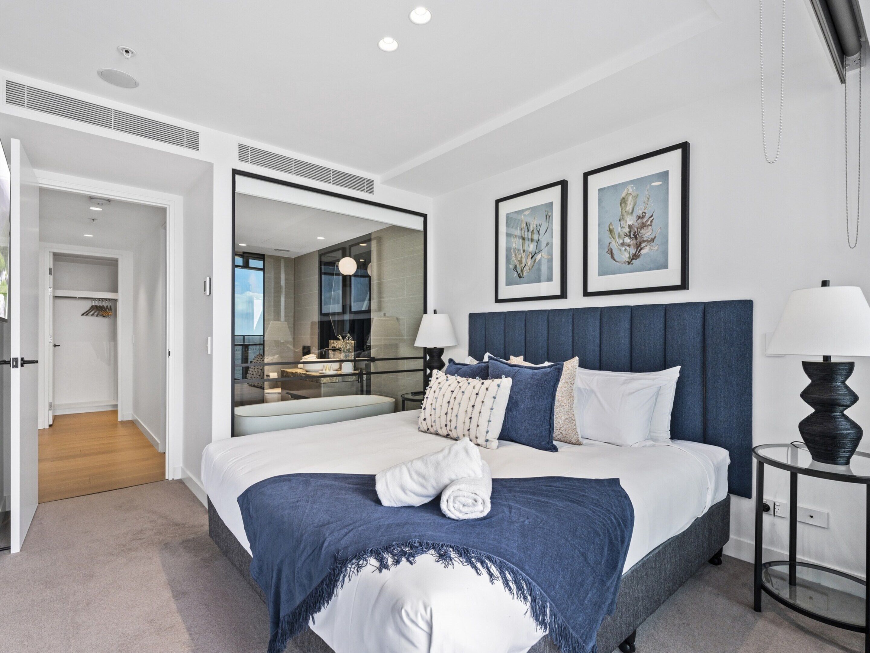 A modern bedroom with a large bed, a bedside table, a mirror, and two framed artworks on the wall.