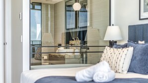 A modern hotel room with a large bed, a glass partition, a view of the city, and a balcony with a chair and table. - Luxury Beachfront 1-bed Apartment With Pool & Gym (Gold Coast)